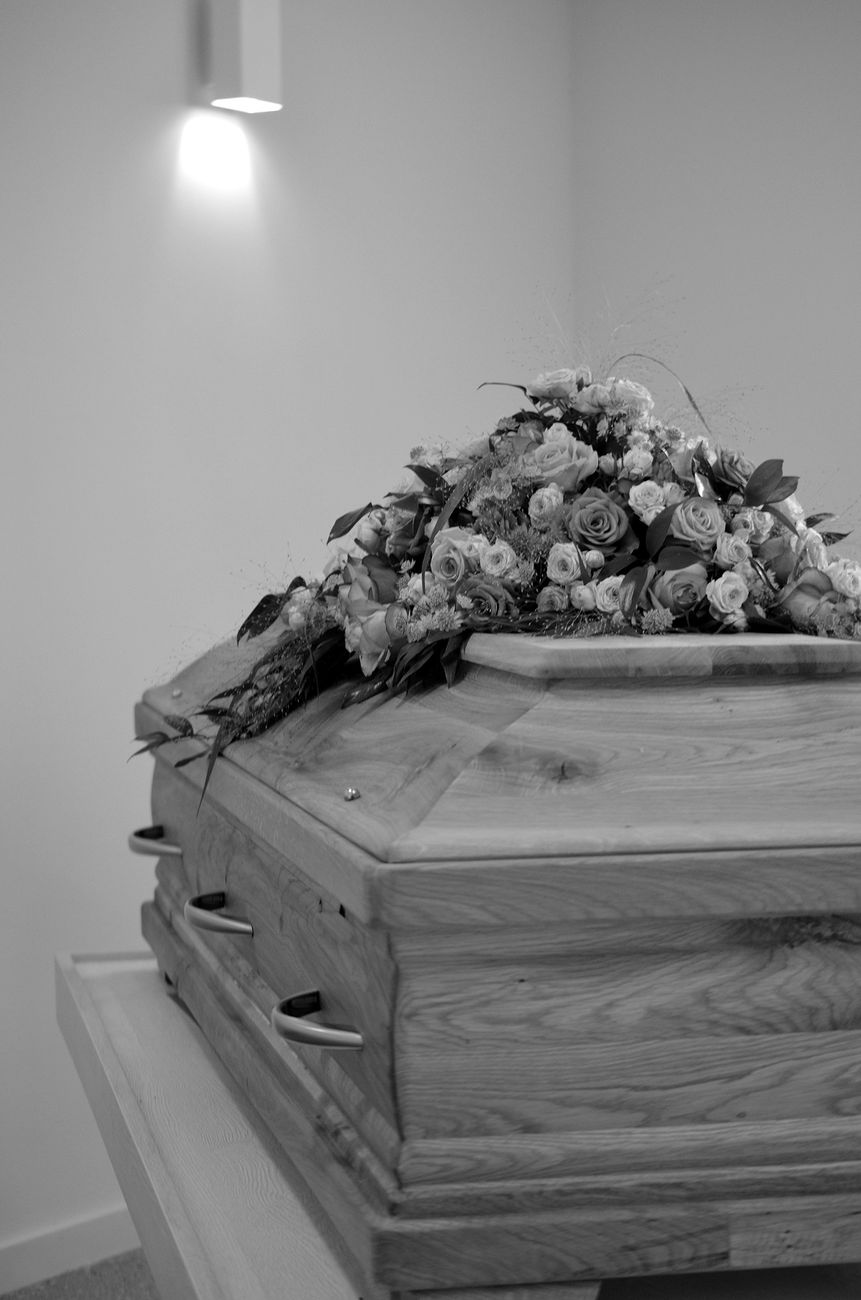 Home A black-and-white image of a wooden casket adorned with flowers in a peaceful setting.